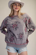 A woman wearing a gray oversized sweatshirt with a pink and green floral print, styled with light blue denim shorts and a white cowboy hat against a plain beige background.