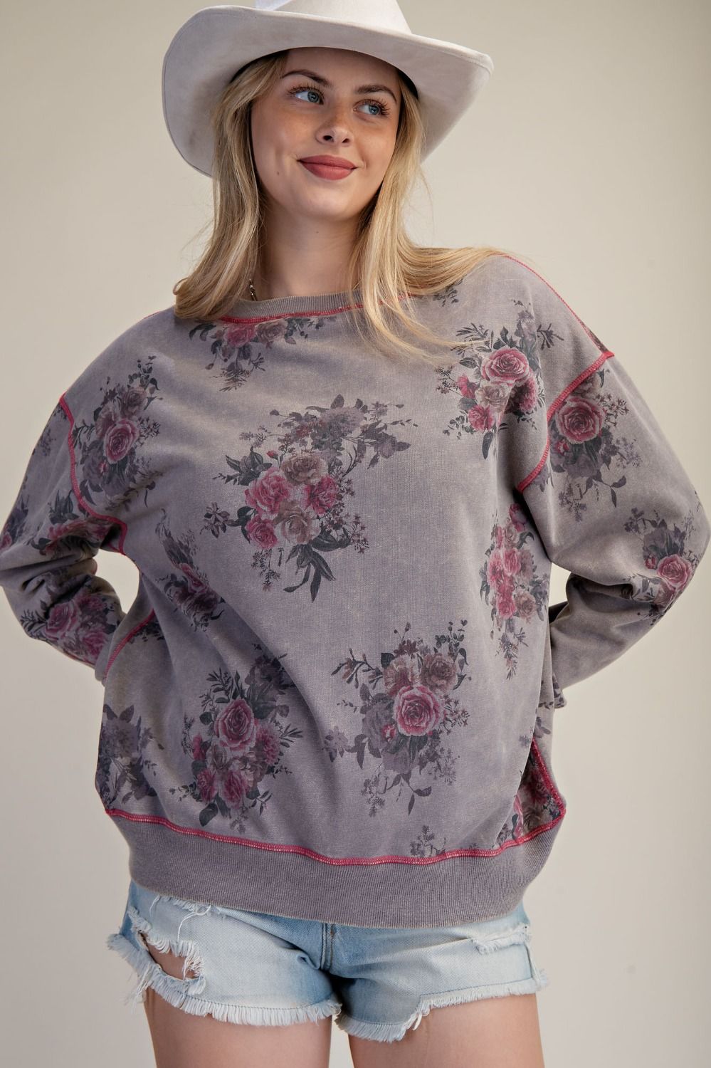 A woman wearing a gray oversized sweatshirt with a pink and green floral print, styled with light blue denim shorts and a white cowboy hat against a plain beige background.