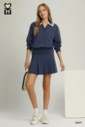 A woman models a navy blue two-piece tracksuit featuring a long-sleeve collared sweatshirt and a matching pleated mini skirt, styled with white sneakers and socks, against a light gray studio background.