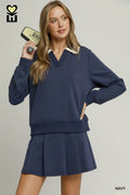 A woman modeling a navy blue long-sleeve sweatshirt and matching pleated mini skirt set. The sweatshirt features a V-neckline with a cream lace trim detail.