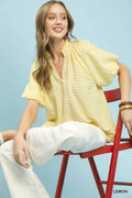 Woman sitting on a red chair wearing a yellow checkered blouse and white pants against a light blue background.