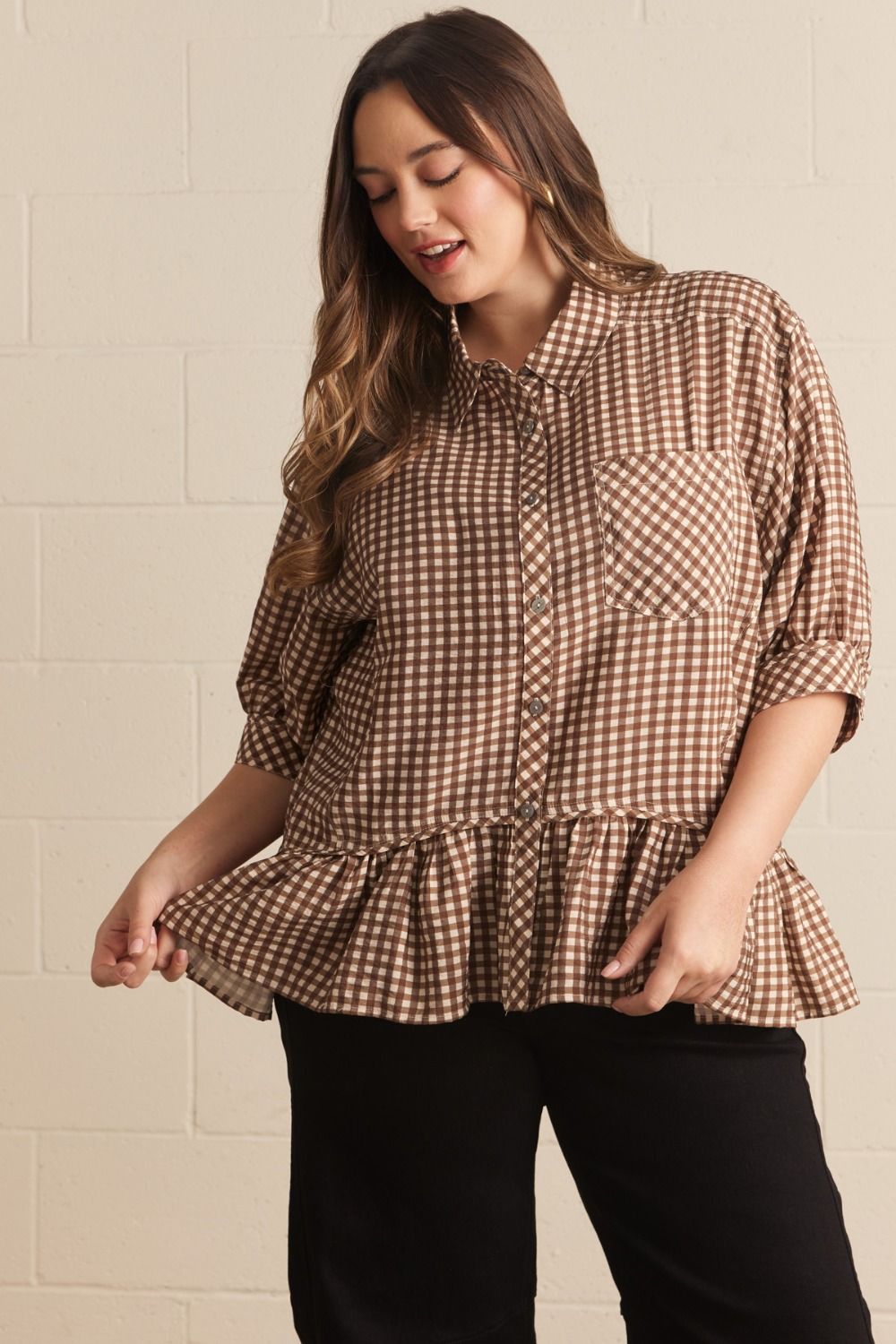 Woman wearing a brown and white checkered blouse with a ruffled bottom against a beige wall.