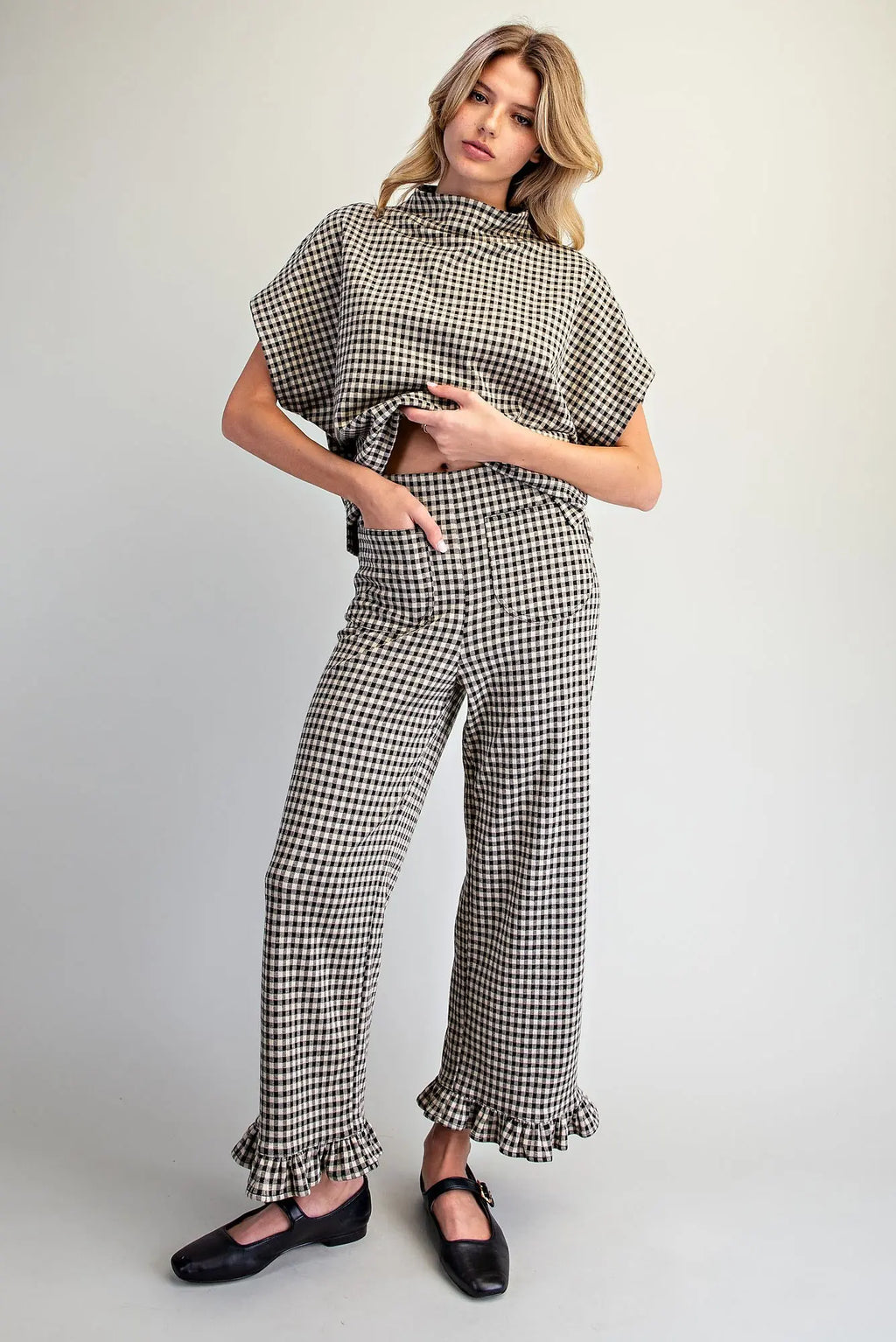 A woman modeling a matching two-piece houndstooth set consisting of a short-sleeve button-front top and wide-leg trousers with ruffled hems, paired with black Mary Jane shoes, against a light gray studio background.