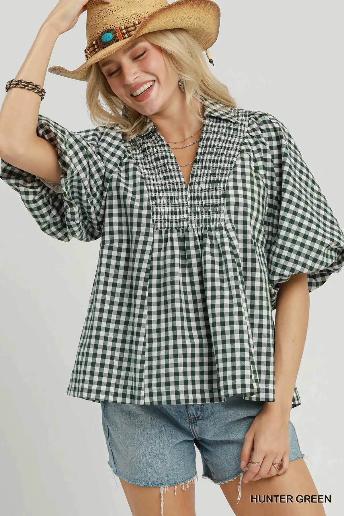 A woman wearing a green and black gingham peplum blouse with short puff sleeves, styled with denim shorts and a straw cowboy hat against a light gray studio background.