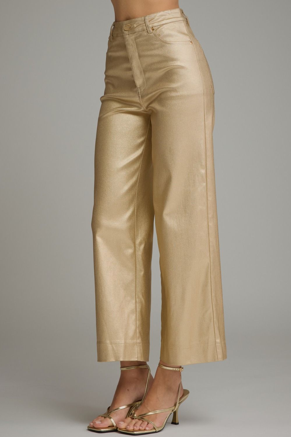 A model is shown from the waist down wearing shiny metallic gold cropped wide-leg pants and matching metallic gold strappy heeled sandals against a solid gray studio background.
