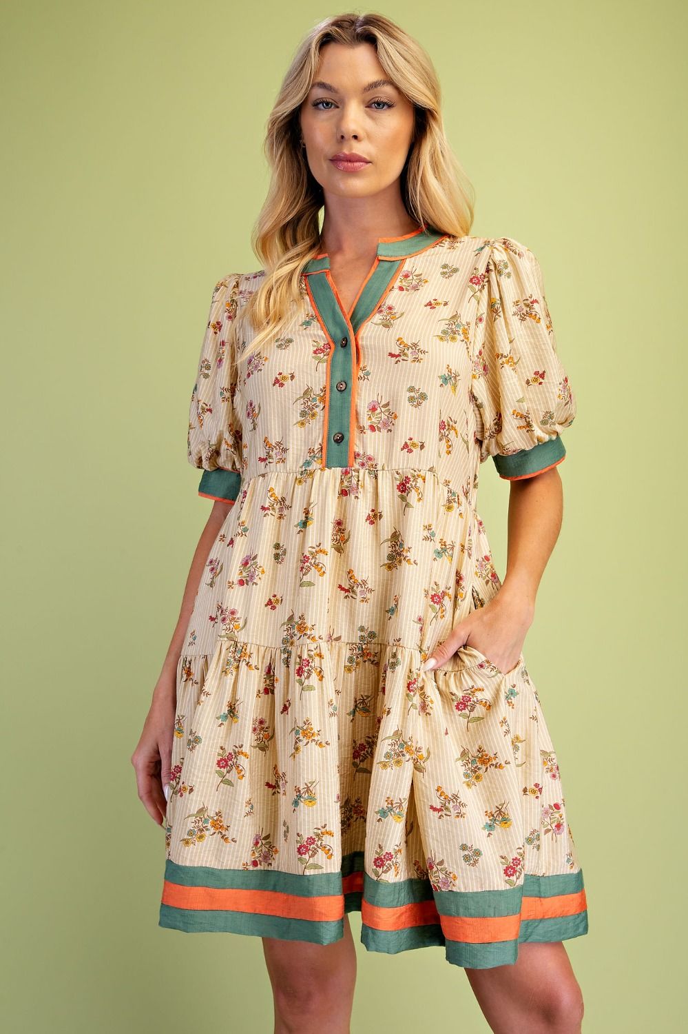 A woman modeling a short-sleeve tiered dress with a beige floral print, teal green trim, and orange button accents, set against a solid light green background.