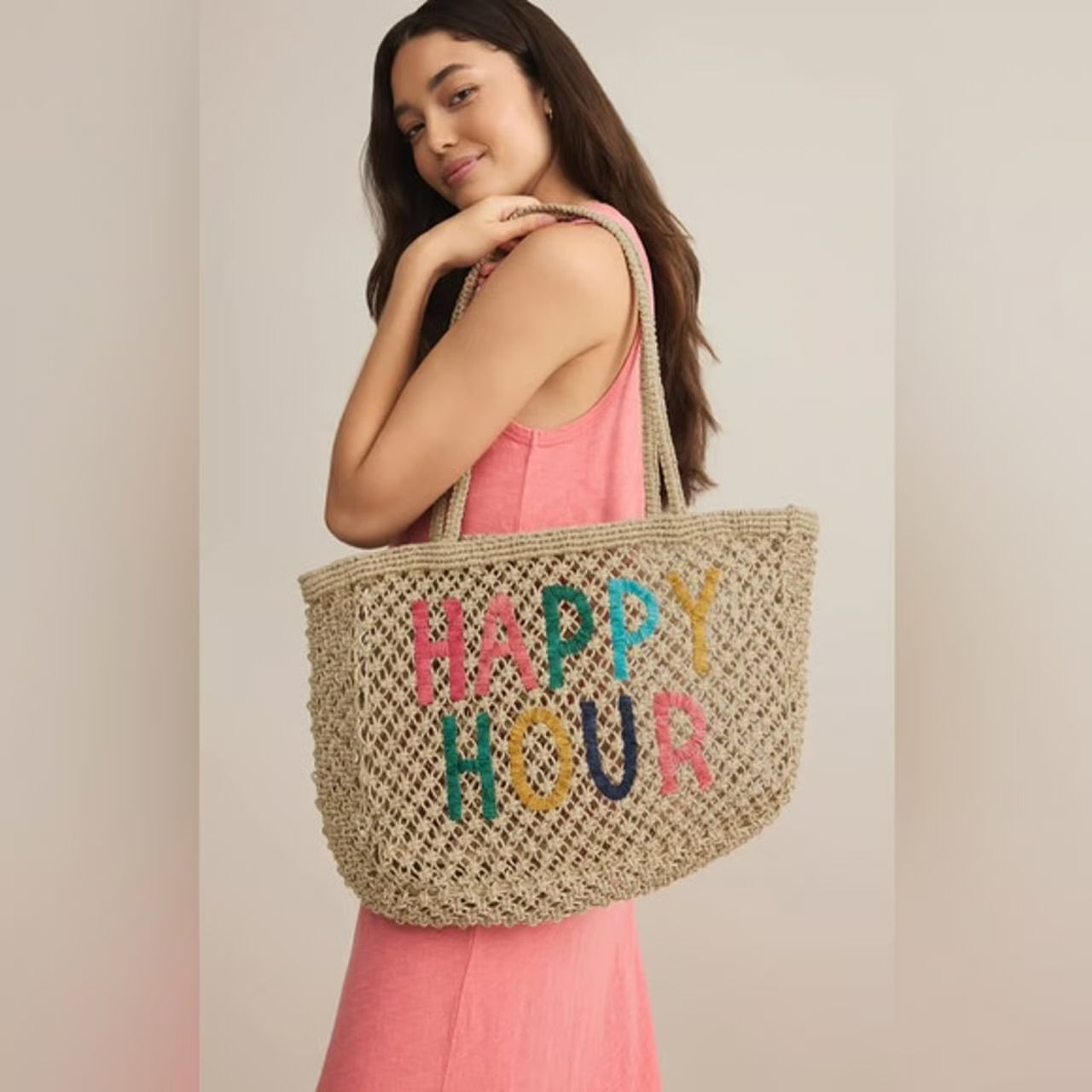 Woman holding a woven bag with 'Happy Hour' text in colorful letters against a plain background