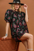 A woman models a short black dress with a vibrant pink and green paisley pattern, styled with a black wide-brim hat and a silver cuff bracelet, sitting on a woven brown chair against a solid brown background.