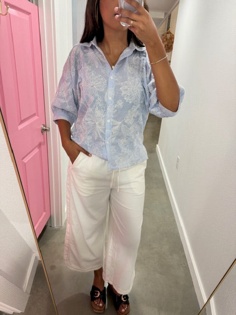 Person taking a mirror selfie wearing a light blue shirt and white pants in a room with a pink door.