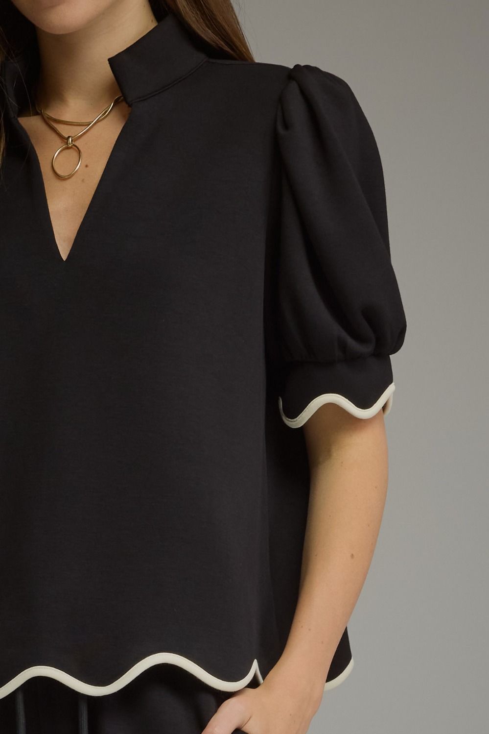 A close-up of a woman wearing a black blouse with a high mock neck, puffed short sleeves, and contrasting white scalloped trim at the sleeve cuffs and hem.