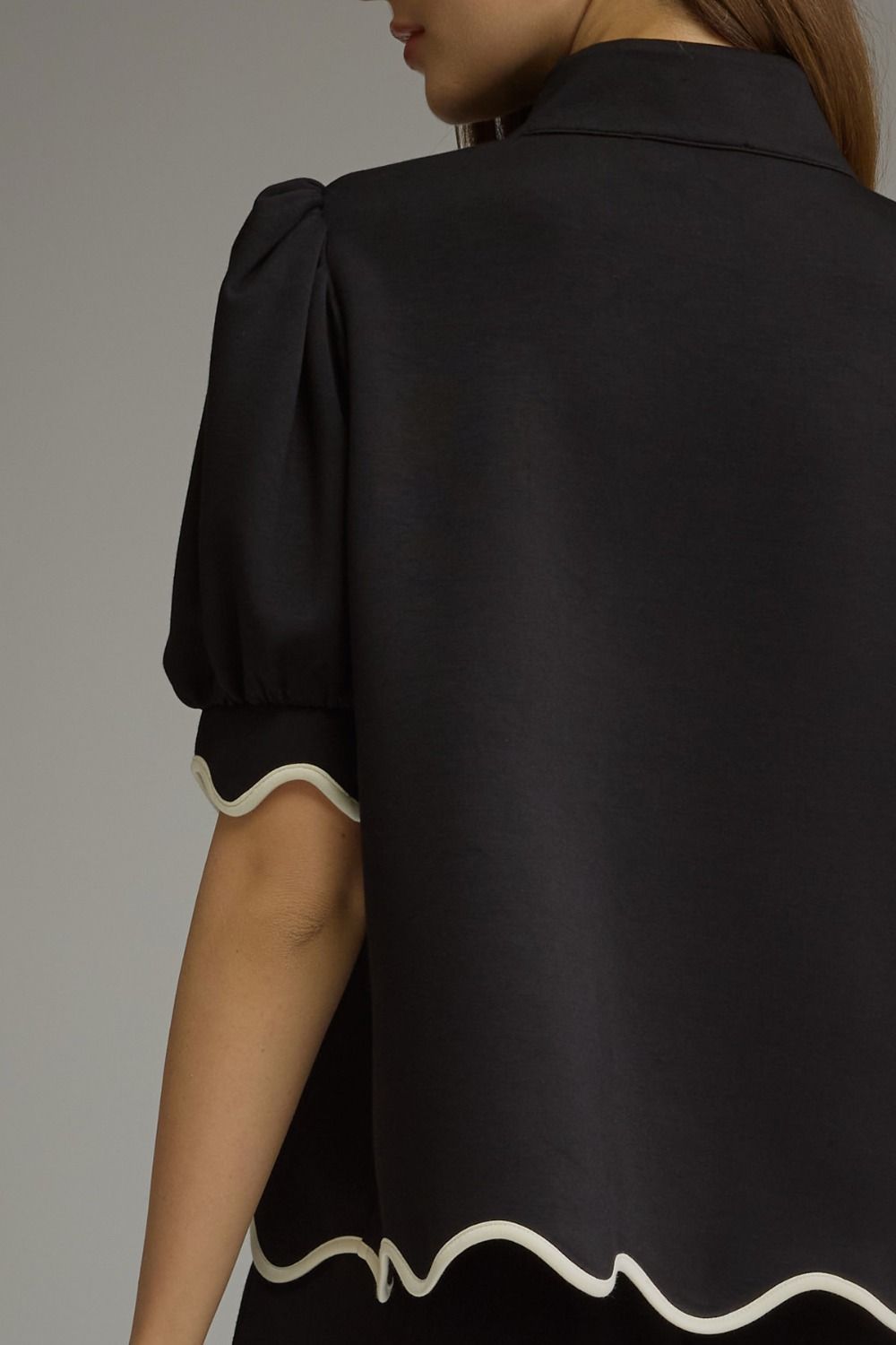 Close-up back view of a black short-sleeve top or cape-style blouse with puff sleeves and a wavy cream trim along the sleeve cuffs and hem, worn by a model against a plain light gray studio background.