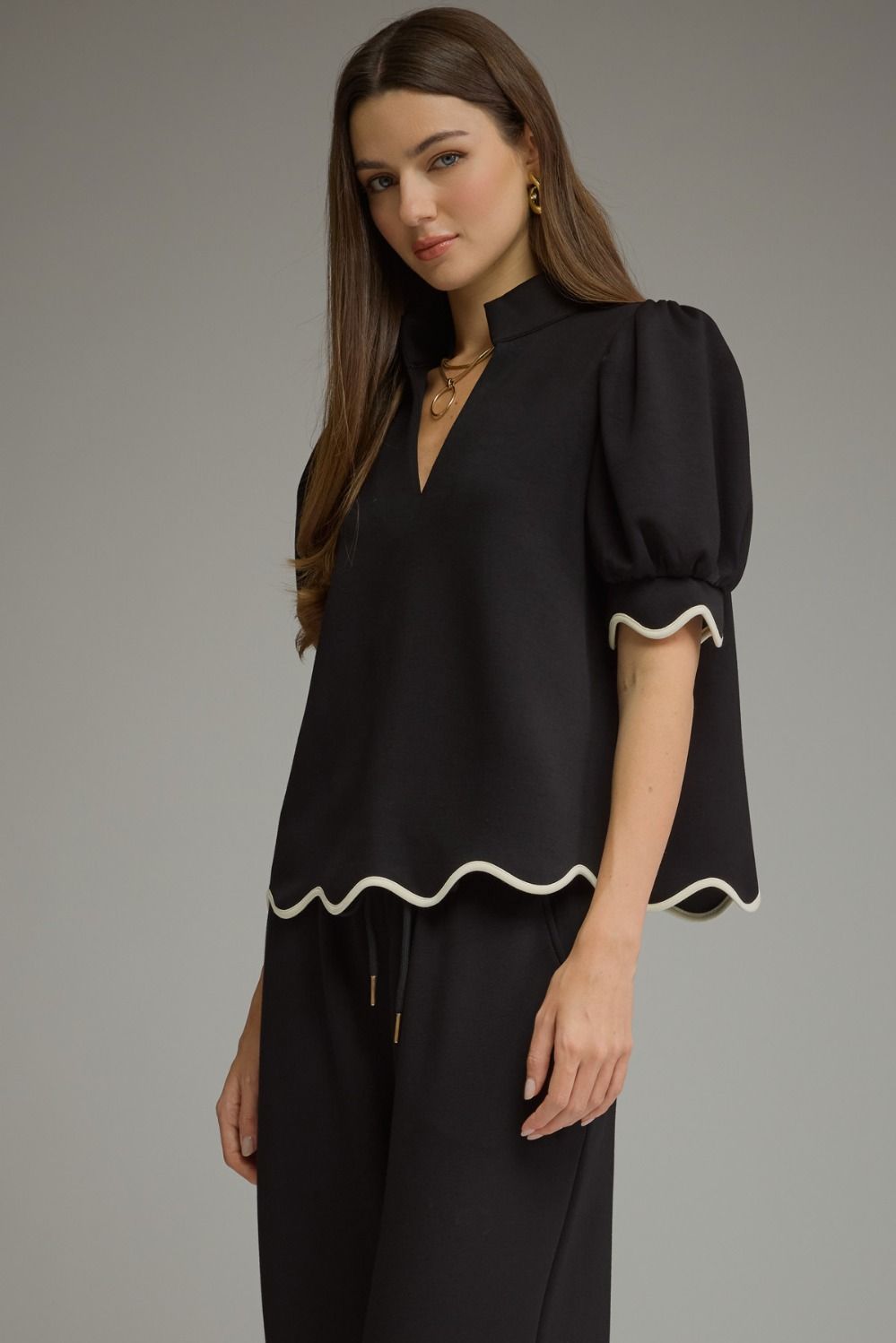 A woman modeling a black blouse with puff sleeves, a high mock neck, and a scalloped white trim, paired with black trousers, against a solid gray studio background.