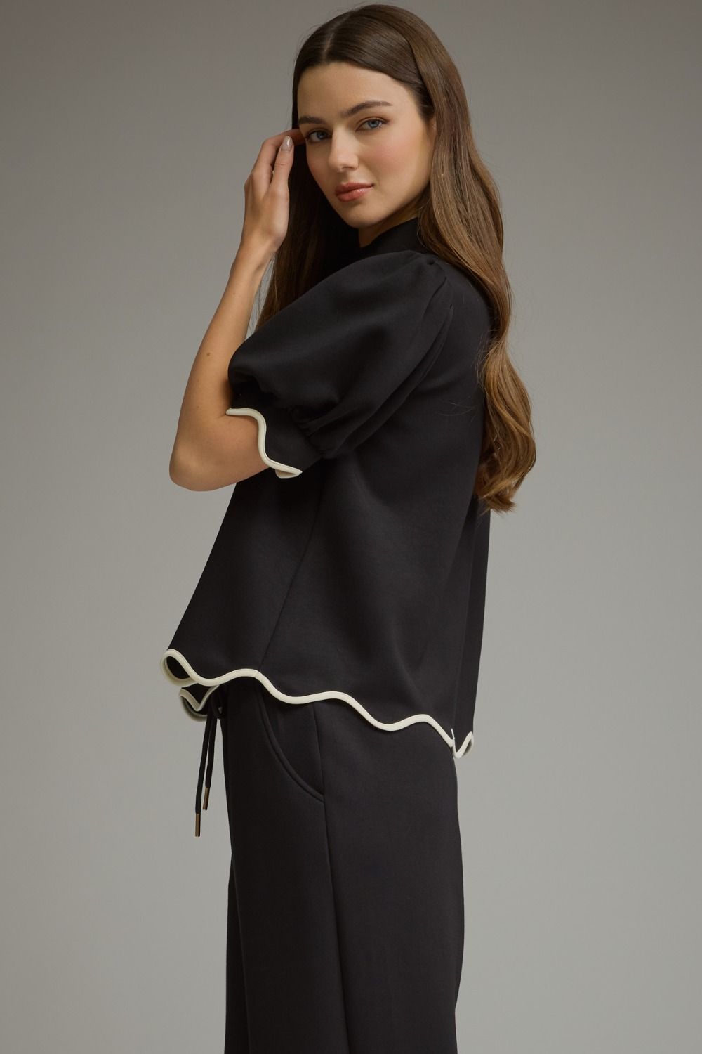 A woman modeling a black short-sleeve top with cream ruffled trim, paired with black wide-leg trousers, against a solid gray studio background.