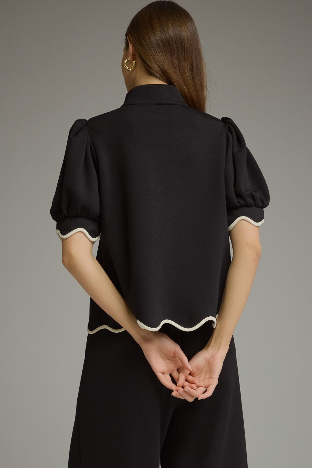 A woman shown from the back wearing a black short-sleeve blouse with cream scalloped trim and matching black trousers, against a plain gray studio background.