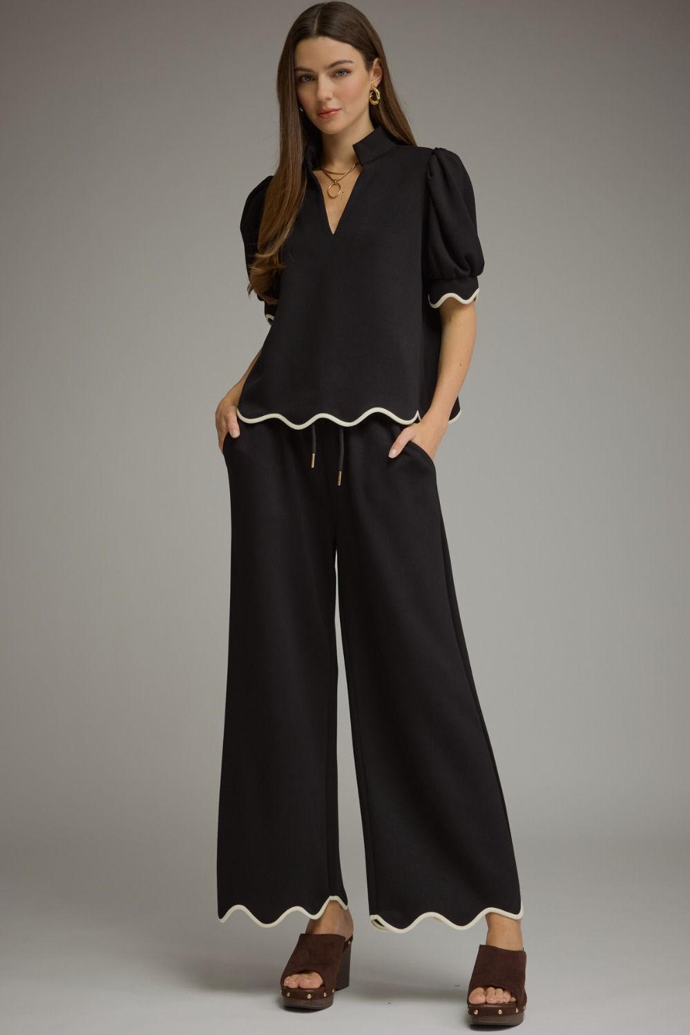 A woman modeling a black set with cream scalloped trim, featuring puff sleeves, a V-neckline, and wide legs, paired with brown wedge sandals against a neutral gray studio background.