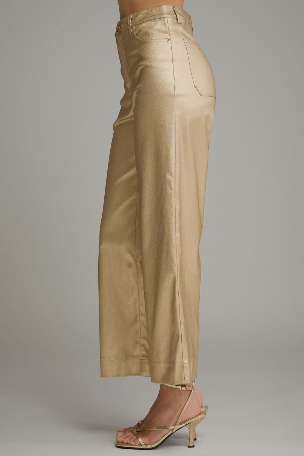A side profile view of a model wearing high-waisted, wide-leg cropped pants in a shiny champagne-gold fabric, paired with matching metallic heeled sandals, against a solid gray studio background.