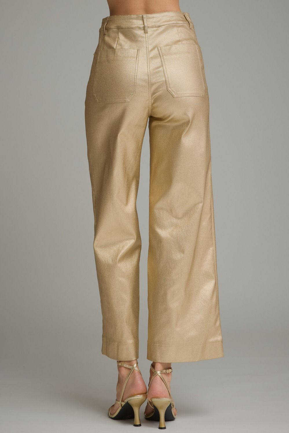 A pair of cropped wide-leg trousers in a shiny metallic gold fabric, shown from the back on a model wearing pink high-heeled sandals against a neutral gray background.