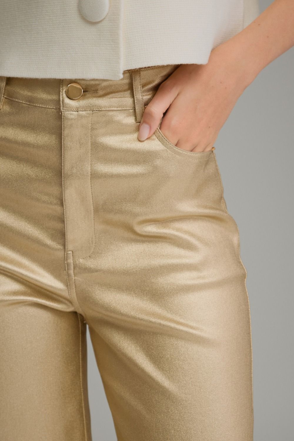 Close-up of a person wearing shiny metallic gold trousers, with a hand in the pocket and part of a cream top visible against a light gray background.