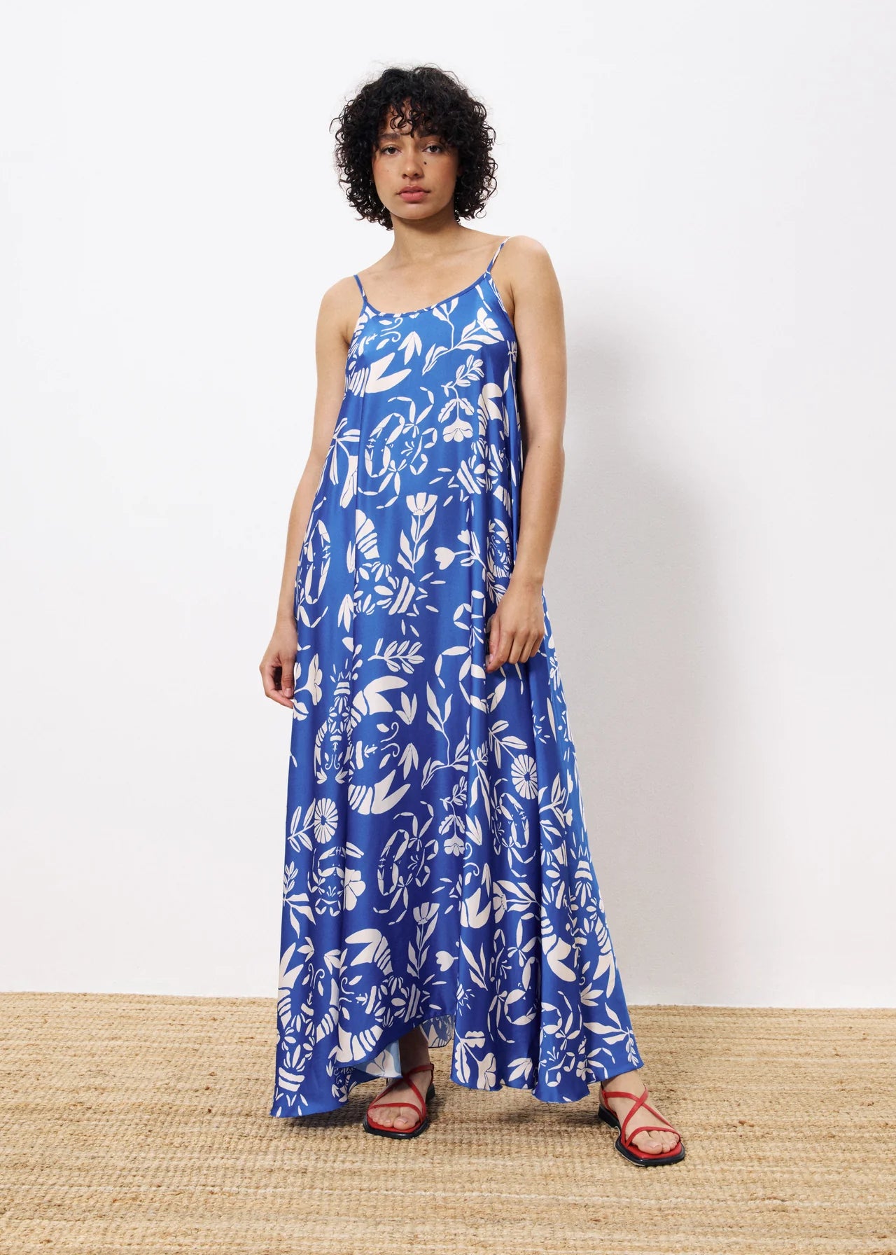 Woman wearing a blue and white floral print maxi dress with spaghetti straps and red sandals, standing against a white background – stylish summer outfit for beach or resort wear