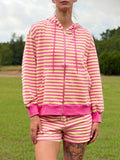 A woman wearing a pink and yellow striped zip-up hoodie and matching striped shorts, standing outdoors in a grassy field under a cloudy sky.