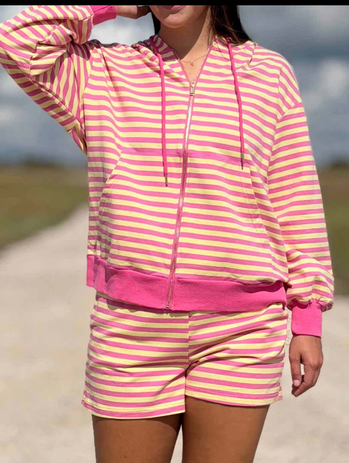 A woman wearing a pink and yellow striped zip-up hoodie and matching shorts, photographed outdoors on a path with a blurred natural background.