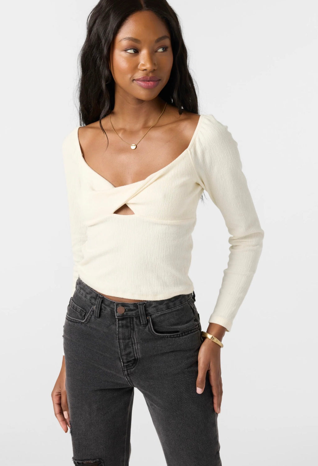 A woman modeling a cream ribbed long-sleeve top with a crossover V-neck and front cutout detail, paired with dark gray jeans, against a plain light gray studio background.