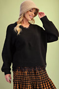 A woman wearing a black knit sweater with a scalloped neckline, a beige bucket hat, and an orange and black plaid skirt, posed against a solid light green background.