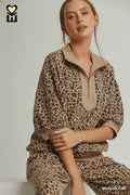 A woman wearing a beige and brown leopard-print lounge set with a half-zip mock neck top and matching pants, posed against a light gray studio background.