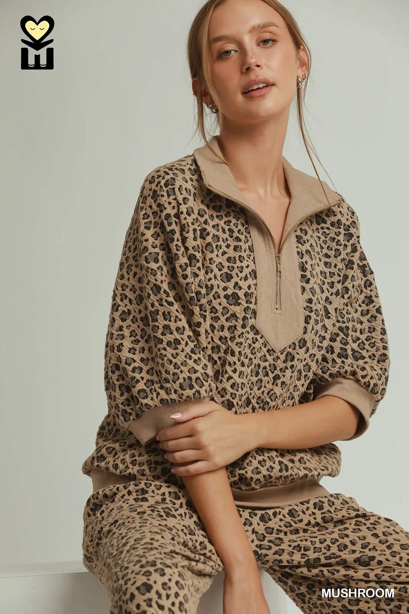 A woman wearing a beige and brown leopard-print lounge set with a half-zip mock neck top and matching pants, posed against a light gray studio background.