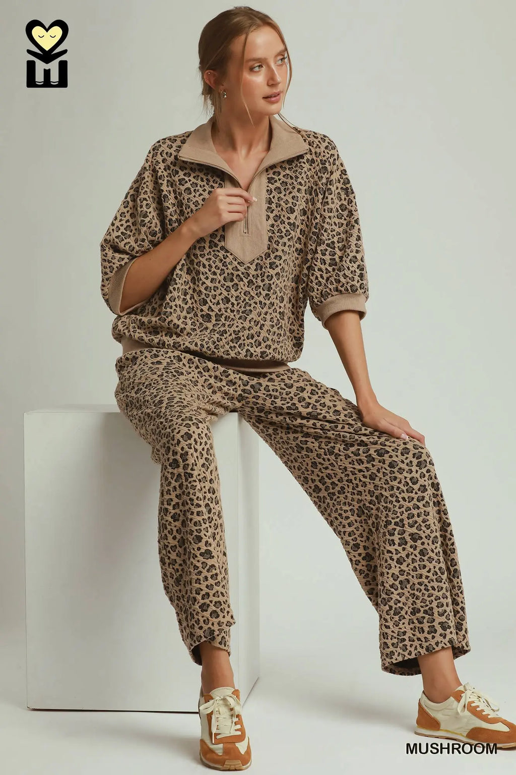 A woman models a matching two-piece lounge set with a brown and beige leopard print pattern, sitting on a white block against a light gray studio background.