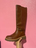 A brown knee-high boot with a ribbed side panel and a buckle strap at the ankle, held up by a hand against a textured pink wall.