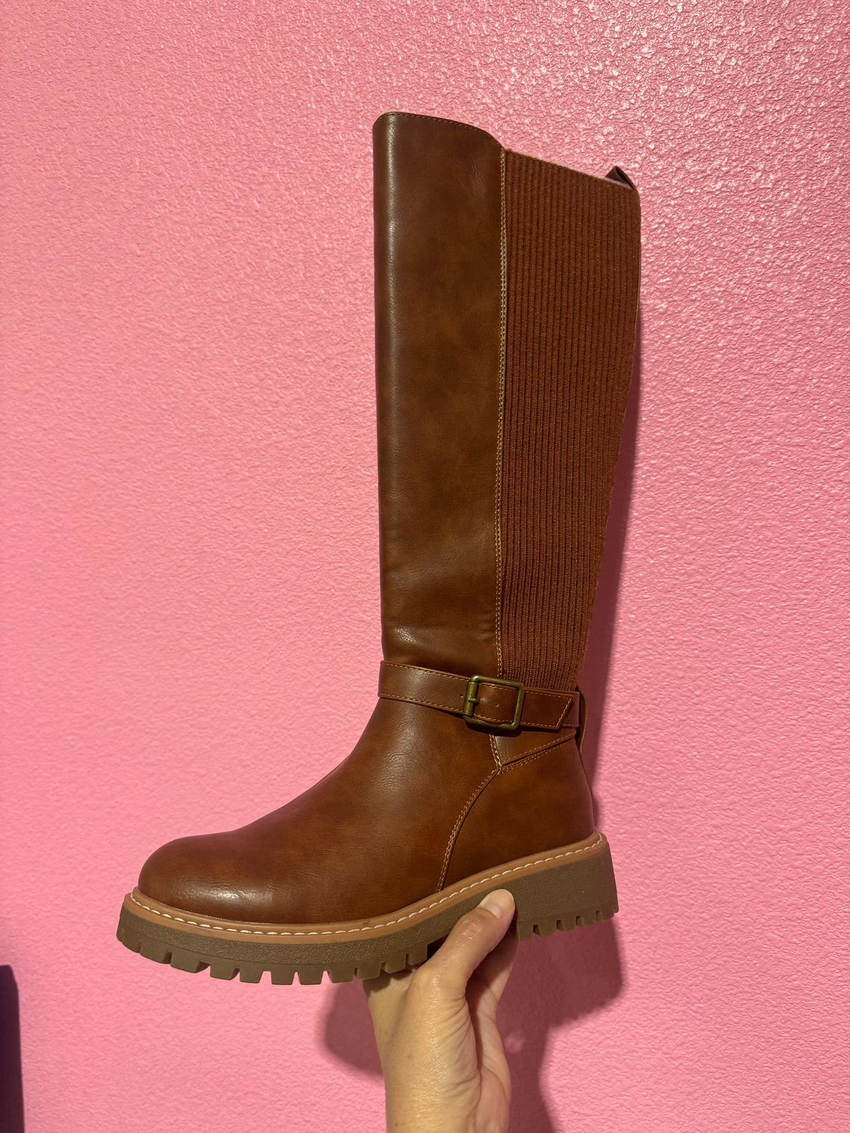 A brown knee-high boot with a ribbed side panel and a buckle strap at the ankle, held up by a hand against a textured pink wall.