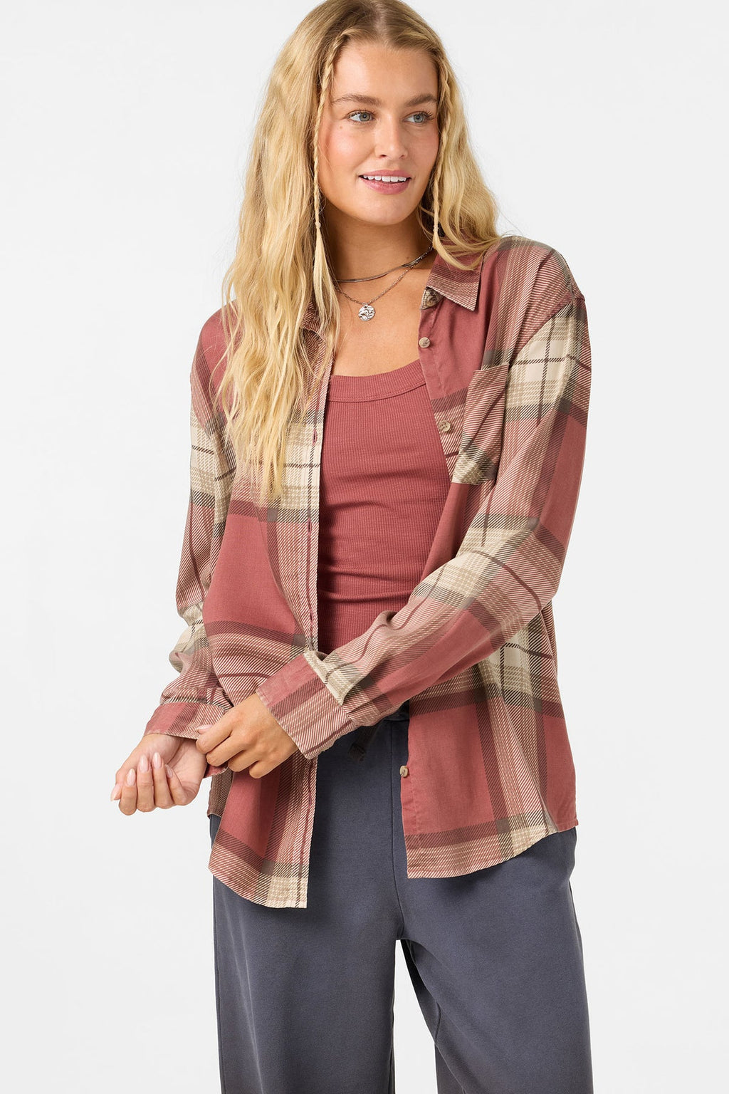 A woman modeling a rust-colored plaid button-up shirt over a dusty rose tank top and dark gray pants, photographed against a light gray studio background.
