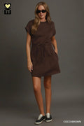 A woman modeling a short-sleeve brown dress with a drawstring waist and side pockets, styled with black sunglasses and black sneakers against a dark gray studio background. The text 'COCO BROWN' appears in the bottom right corner.