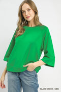 A woman wearing a bright green three-quarter sleeve top and blue jeans, standing against a light gray studio background. Text in the bottom right corner reads 'ISLAND GREEN - UMG'.