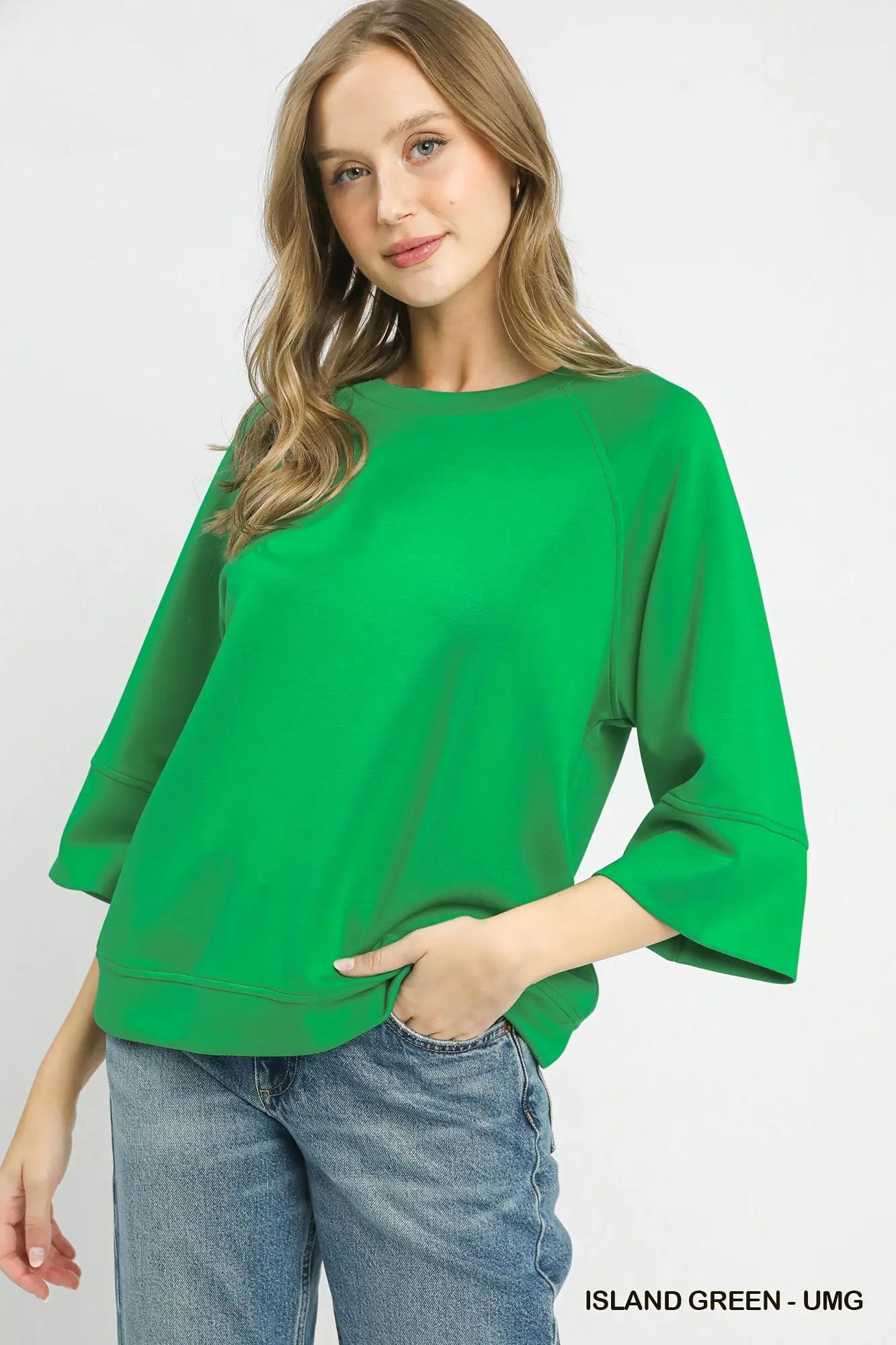A woman wearing a bright green three-quarter sleeve top and blue jeans, standing against a light gray studio background. Text in the bottom right corner reads 'ISLAND GREEN - UMG'.
