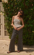 Woman wearing a polka dot strapless top and wide-leg pants standing against a green leafy background.