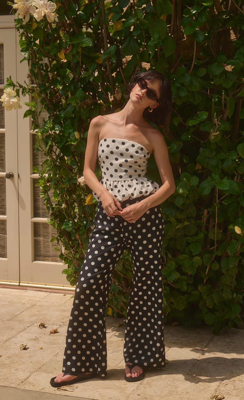 Woman wearing a polka dot strapless top and wide-leg pants standing against a green leafy background.