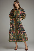 A woman modeling a long-sleeved midi shirt dress with a colorful floral and paisley print on a dark background, featuring a tie waist belt and pointed shoes.