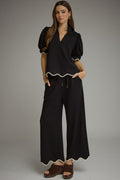 A woman modeling a black two-piece outfit consisting of a short-sleeve top with puff sleeves and a wavy hem, paired with matching wide-leg pants featuring a wavy hem and drawstring waist, styled with brown wedge sandals against a gray studio background.