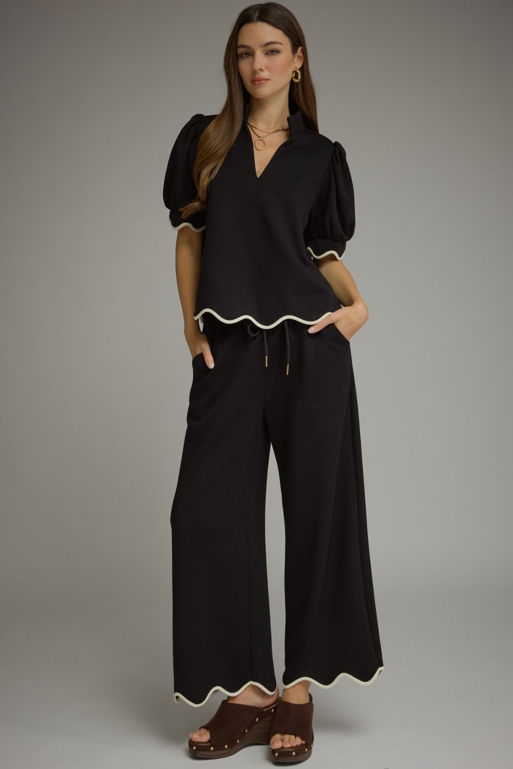 A woman modeling a black two-piece outfit consisting of a short-sleeve top with puff sleeves and a wavy hem, paired with matching wide-leg pants featuring a wavy hem and drawstring waist, styled with brown wedge sandals against a gray studio background.