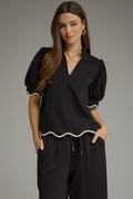 A woman modeling a black short-sleeve blouse with puff sleeves and a wavy white trim, paired with black pants, against a solid gray studio background.