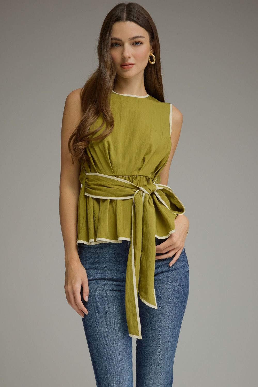 A woman modeling a sleeveless olive green wrap-style top with a tie waist, paired with blue jeans, against a plain gray studio background.