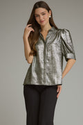 A woman modeling a metallic silver short-sleeve blouse with puffed shoulders and a stand collar, paired with black pants, against a solid gray studio background.