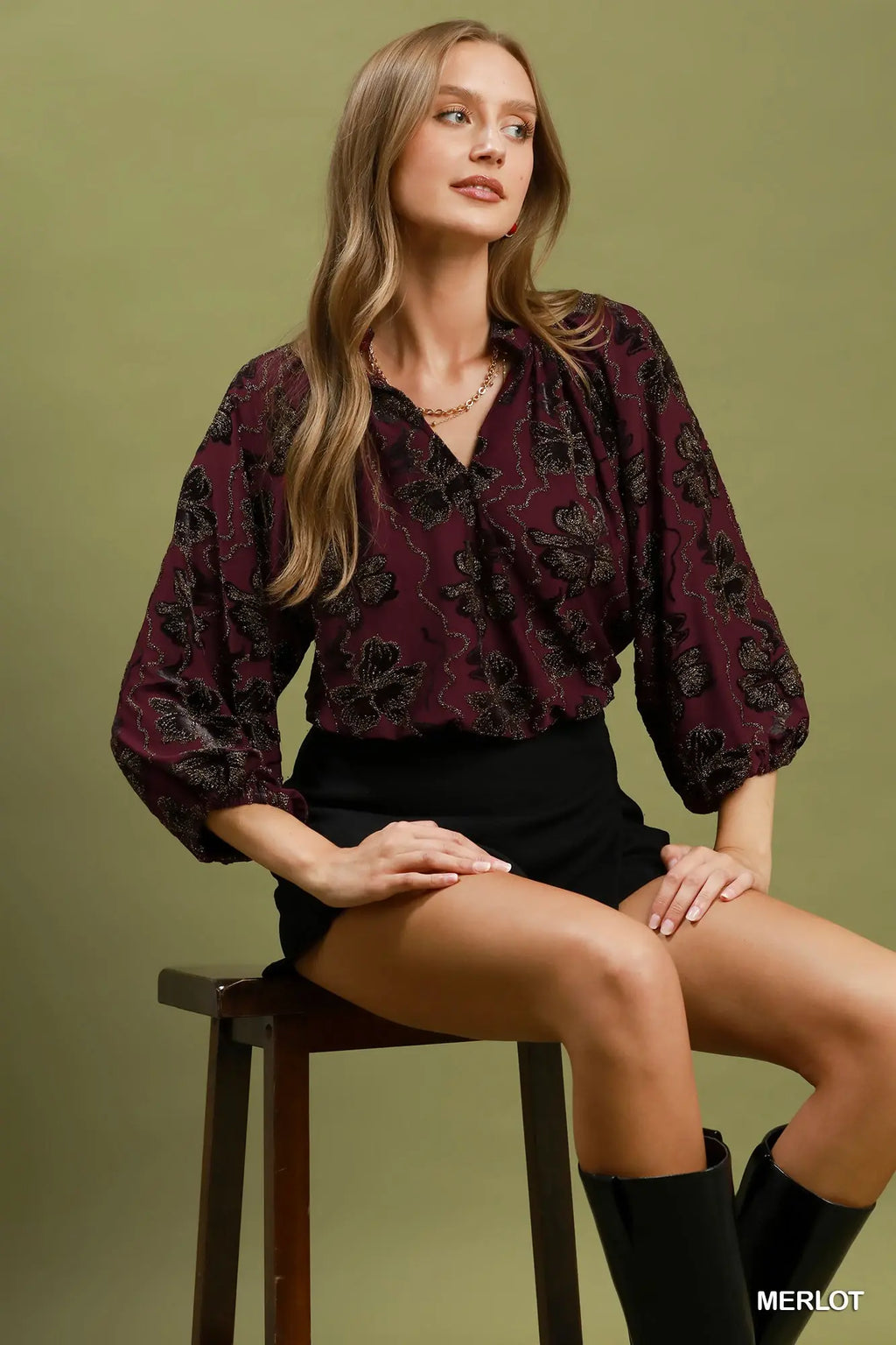 Woman sitting on a stool wearing a burgundy patterned blouse with a green background