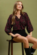 Woman sitting on a stool wearing a burgundy patterned blouse with a green background