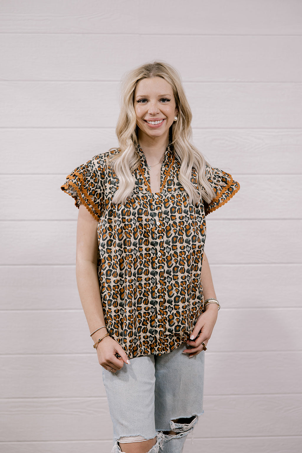A woman with blonde hair wearing a brown and beige leopard print short-sleeve blouse with ruffled trim, paired with light-wash ripped jeans, standing against a white wood-paneled wall.