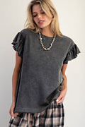 A woman wearing a dark gray washed short-sleeve t-shirt with ruffled sleeves and a plaid skirt, accessorized with a beaded necklace, against a plain light gray background.