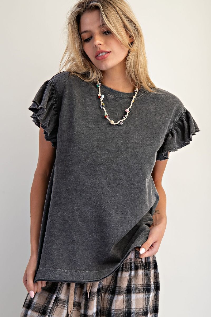 A woman wearing a dark gray washed short-sleeve t-shirt with ruffled sleeves and a plaid skirt, accessorized with a beaded necklace, against a plain light gray background.