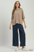 A woman modeling a loose-fit, short-sleeve knit top with horizontal mocha brown and cream stripes, paired with dark navy wide-leg trousers and white sneakers, against a light gray studio background.